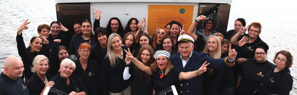 Eine Gruppe von Menschen, einige in Marineuniformen und Matrosenhüten, lächeln und posieren für ein Gruppenfoto am Wasser, halten Friedenszeichen hoch und winken in die Kamera.