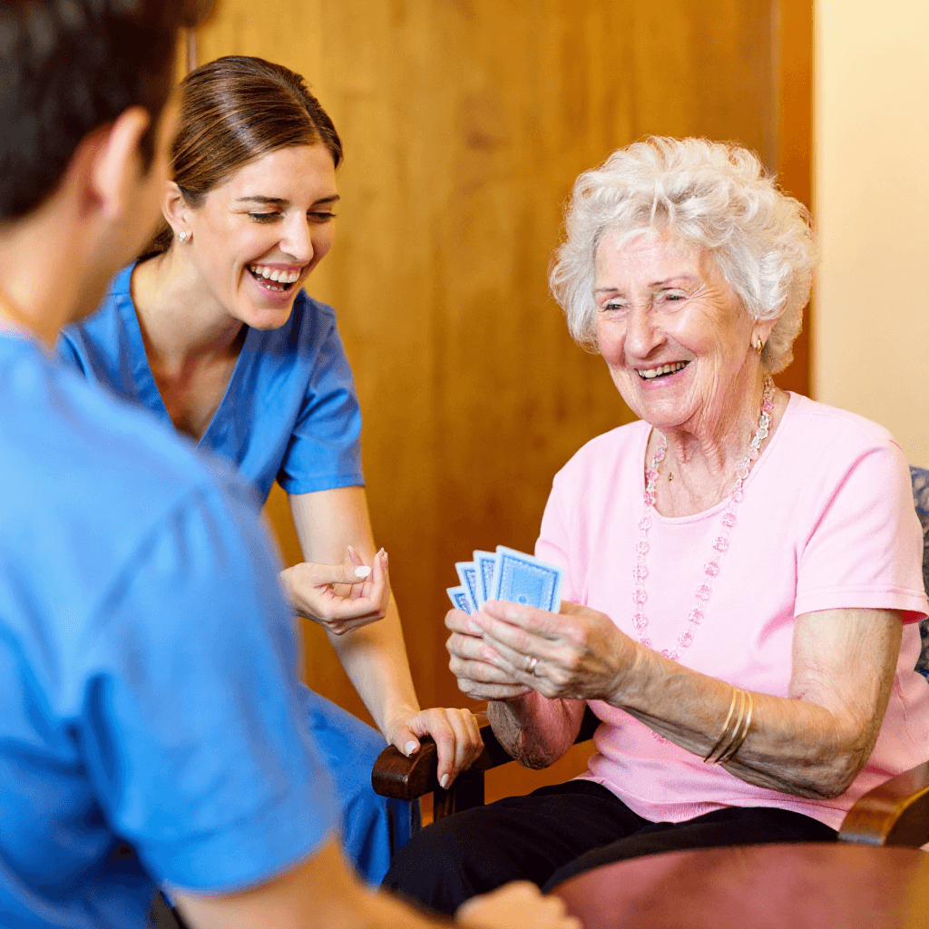 Eine ältere Frau lächelt, während sie Spielkarten in der Hand hält und neben einem Pfleger in blauem Kittel und einer weiteren Person sitzt. Sie alle scheinen ein gemeinsames Spiel in einem hellen, gemütlichen Raum zu genießen.